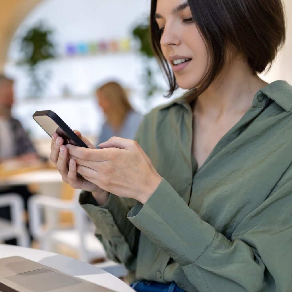 Concentrated young female office worker seated at the laptop screen looking at the smartphone touchscreen